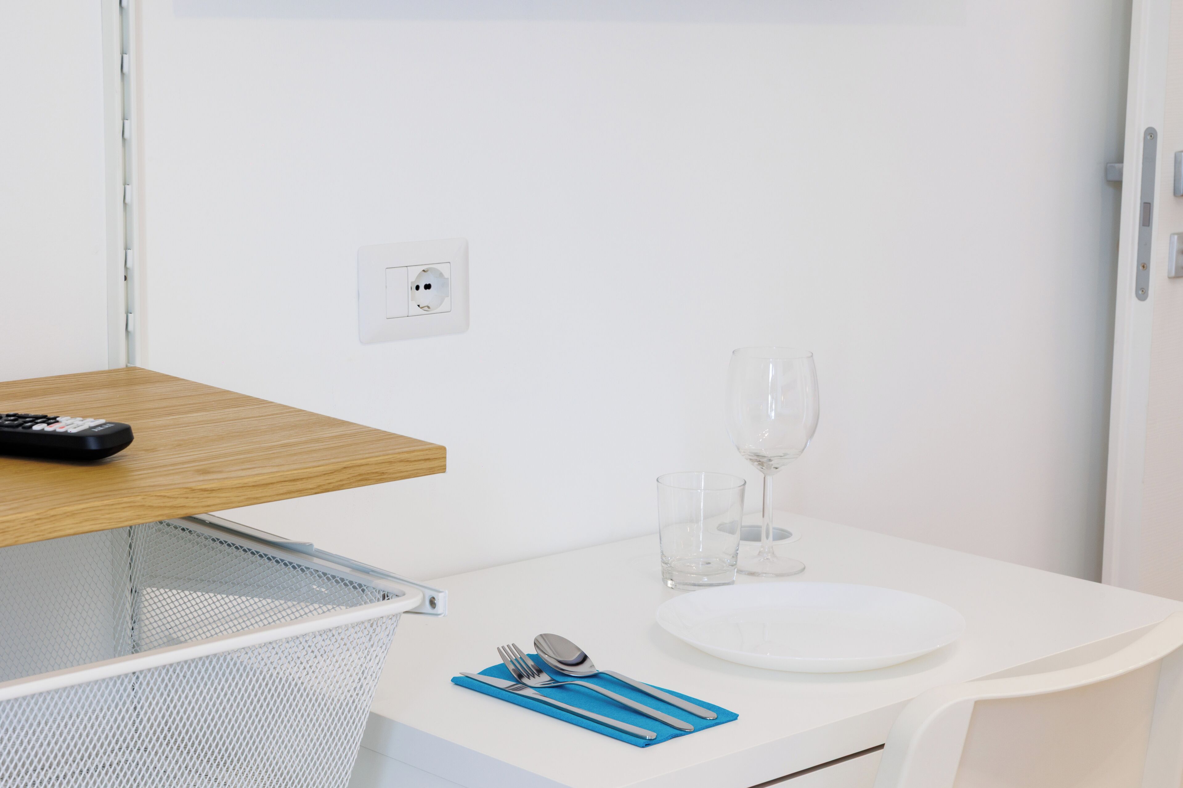 A clean, white room with a wooden shelf, a white table with a plate, cutlery, and a glass, and a power outlet on the wall.