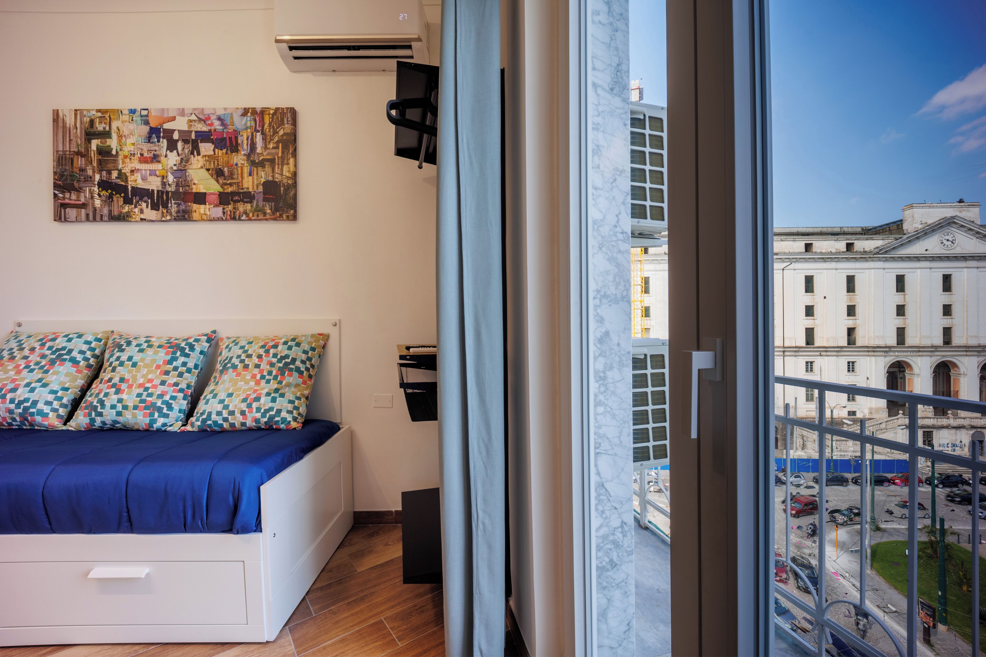 A bedroom with a bed, a wall-mounted air conditioner, a painting, and a view of a building through a window.