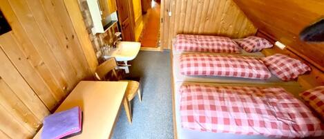 Una cabaña con interior de madera, una cama individual con ropa de cama a cuadros, una mesa pequeña con una silla, y un baño con lavamanos y espejo.