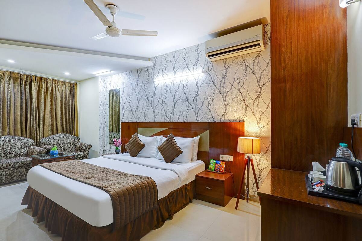 A hotel room with a bed, a desk, a chair, a ceiling fan, an air conditioning unit, and a nightstand with a lamp and a tray of items.