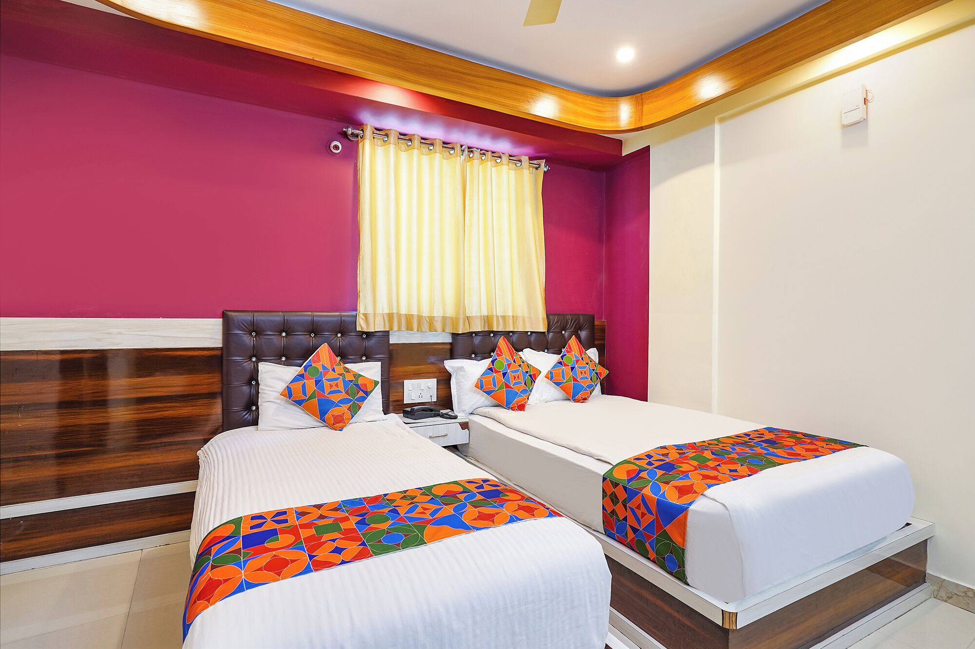 A hotel room with two beds, a wooden headboard, a red accent wall, and a window with yellow curtains.