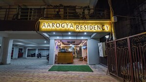 The exterior of AAROGYA RESIDENCY with a reception desk, a gated entrance, and a parking area with scooters.