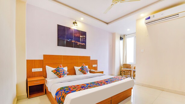 A bedroom with a wooden bed, white bedding, a colorful geometric throw, a nightstand, a wall-mounted lamp, a ceiling fan, and an air conditioning unit.