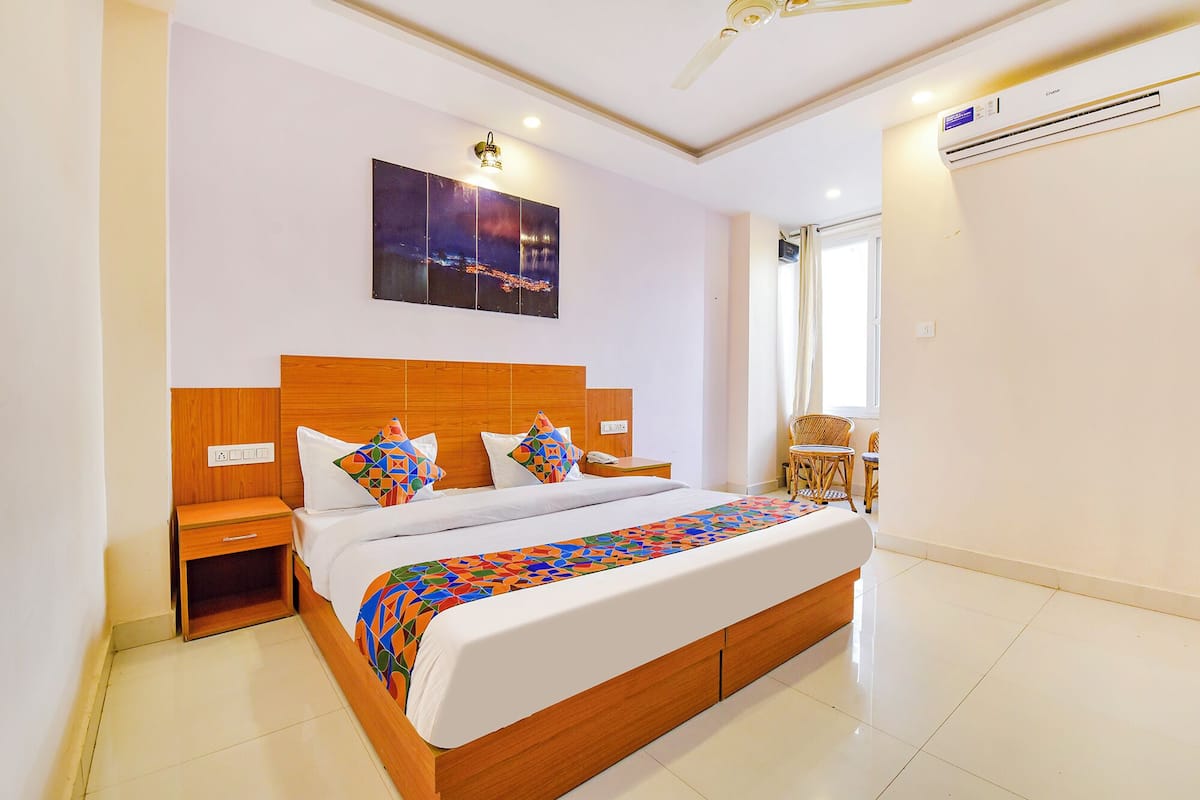 A bedroom with a wooden bed, white bedding, a colorful geometric throw, a nightstand, a wall-mounted lamp, a ceiling fan, and an air conditioning unit.