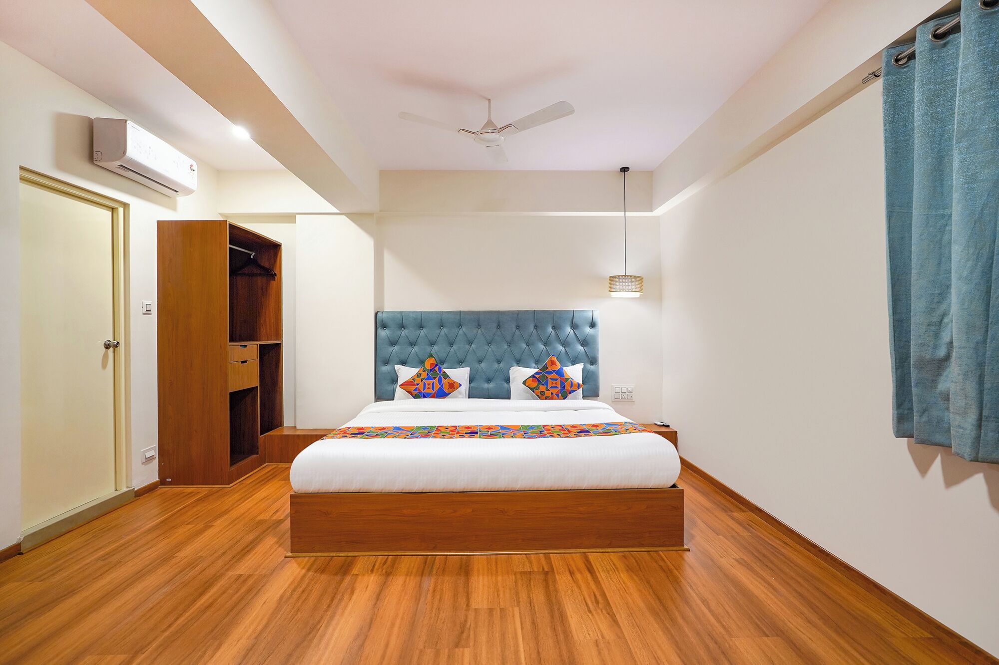 A bedroom with a tufted headboard, a bed with a colorful quilt, a wooden wardrobe, and a ceiling fan.