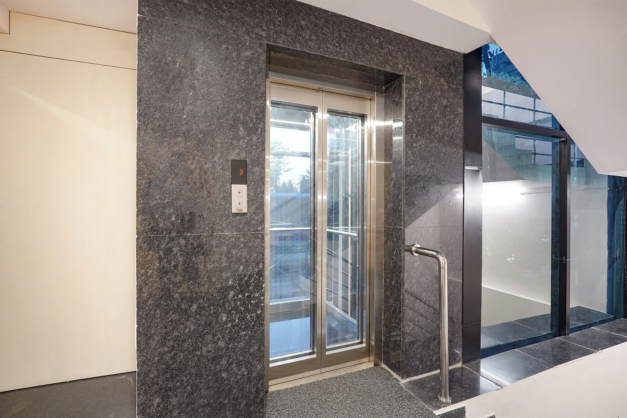 An elevator with a glass door, a metal handrail, and a control panel with an emergency button.