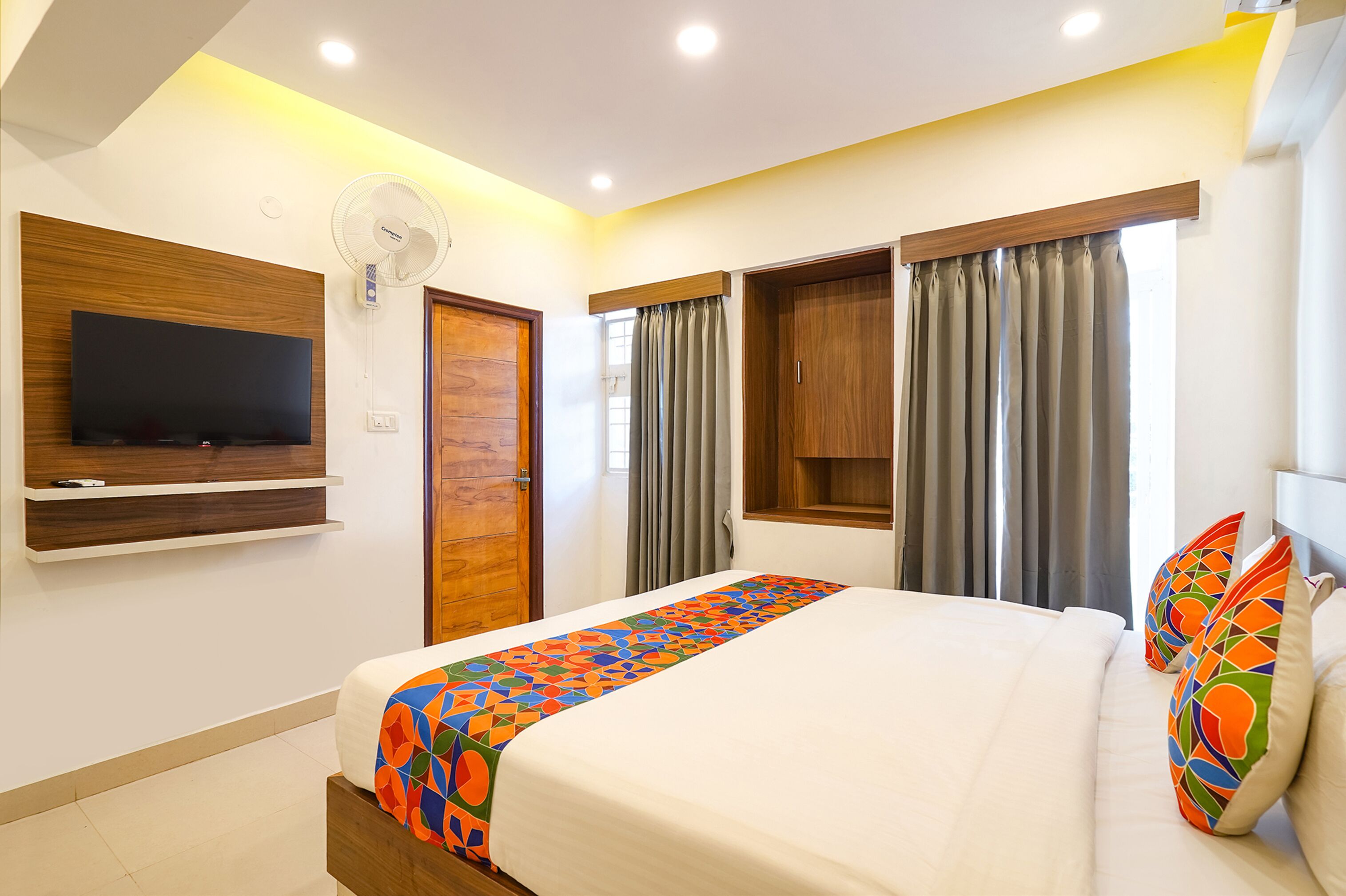 Una habitación de hotel con cama, televisión montada en la pared, ventana con cortinas y ventilador de techo.