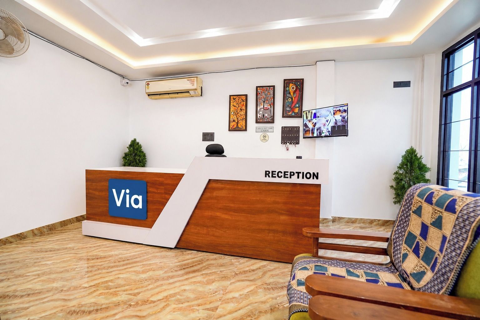 A reception area with a wooden reception desk labeled 'Via' and a reception sign. There are framed artworks on the wall, a television, and a potted plant.