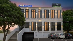 A two-story building with a sign for 'fabHOTELS' and 'The Oak Tree' restaurant. There are three cars parked in front, and a staircase leading to the entrance. - Fabhotel The Oak Tree (Faizabad)