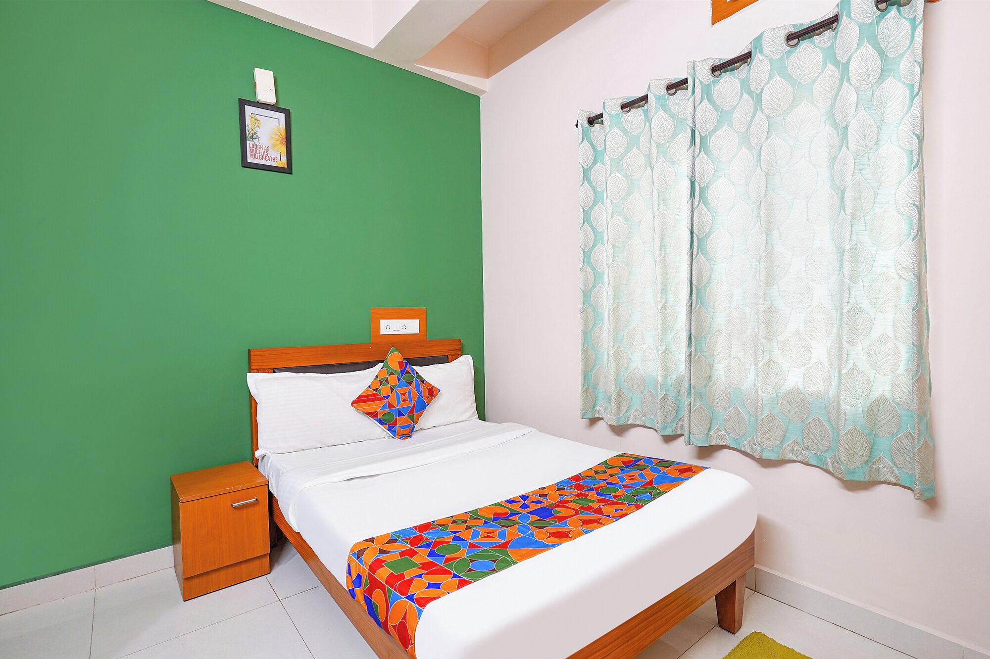 A bedroom with a green wall, a wooden bed frame, a white bedspread, a colorful geometric pillow, a small wooden nightstand, and a window with a leaf-patterned curtain.