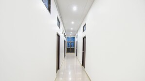 A long, narrow hallway with white walls and a tiled floor, leading to a room with a large window and a door. - Fabhotel Jayaditya Palace (Ayodhya)