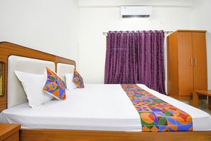 Cama bem arrumada com colchão branco e almofadas geométricas coloridas, cabeceira de madeira, cortina roxa e guarda-roupa de madeira.