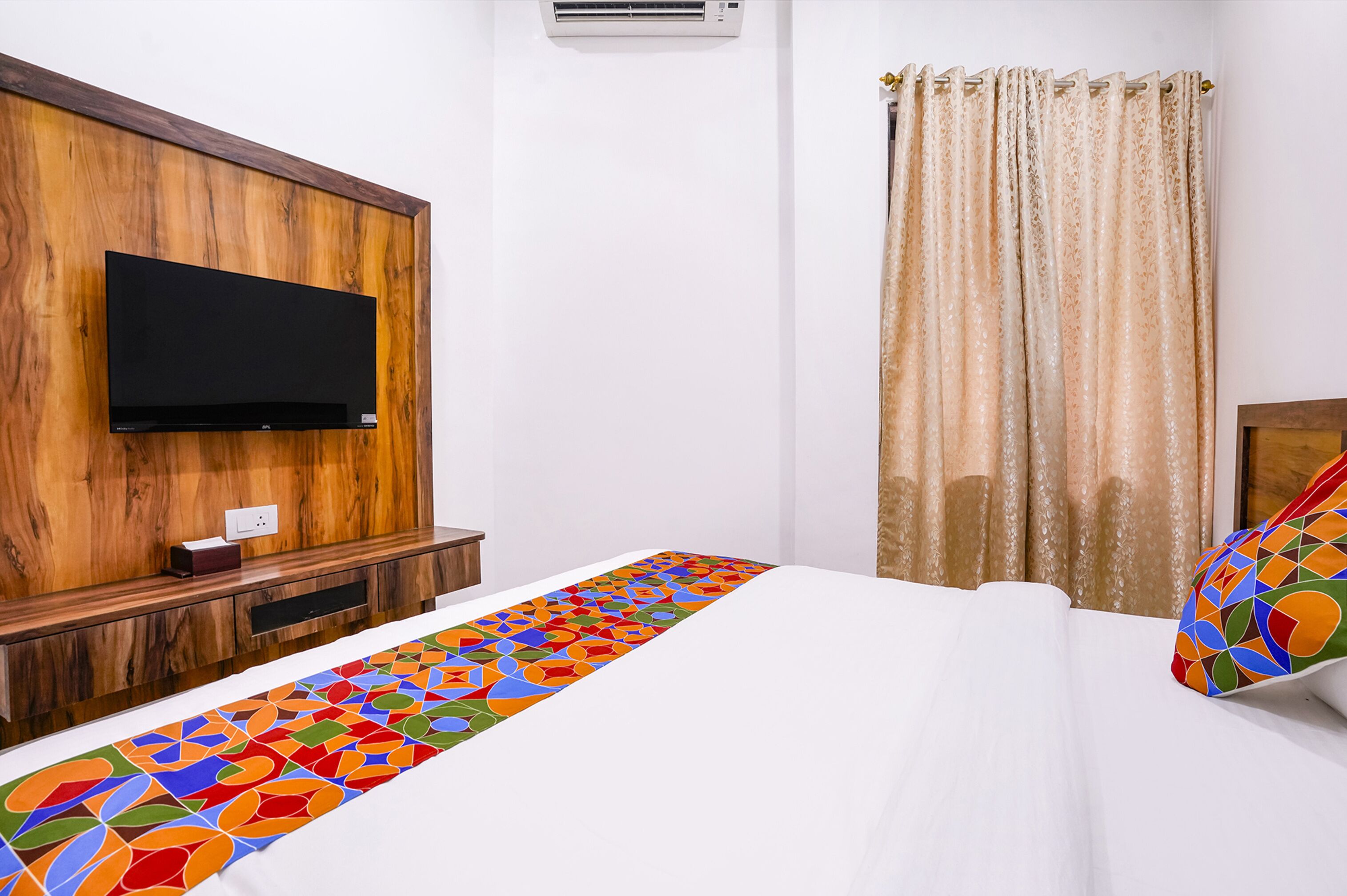 A hotel room with a wooden headboard, a flat-screen TV mounted above it, a bedside table with a lamp, and a colorful runner on the bed.