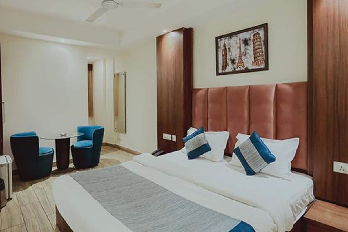 A hotel room with a large bed, two blue chairs, a small table, and a painting of Paris landmarks on the wall.