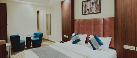 A hotel room with a large bed, two blue chairs, a small table, and a painting of Paris landmarks on the wall.