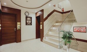 Eine Hotelhalle mit Marmorboden, Holztüren, einer Uhr, einer Treppe mit Glastrennstäben und einem Blumentopf.