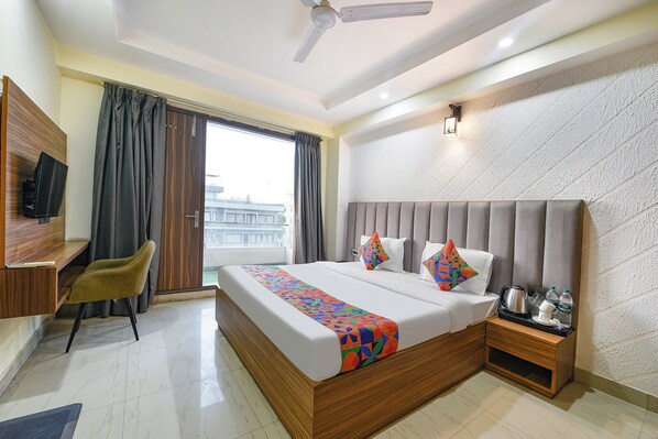 A hotel room with a bed, a desk with a TV, a chair, a nightstand with a kettle, and a view of the city through the window. - Fabhotel Dm 45 (Gurugram)