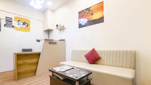 A reception area with a reception desk, a sofa with a red pillow, a coffee table with magazines, and a painting of a sunset on the wall. - Via 24/7 (Navi Mumbai)