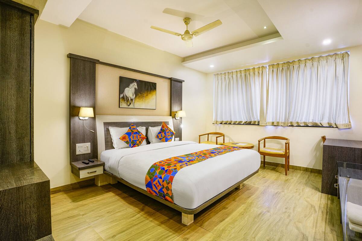 A hotel room with a large bed, a ceiling fan, a painting on the wall, a window with curtains, and a desk with a chair.