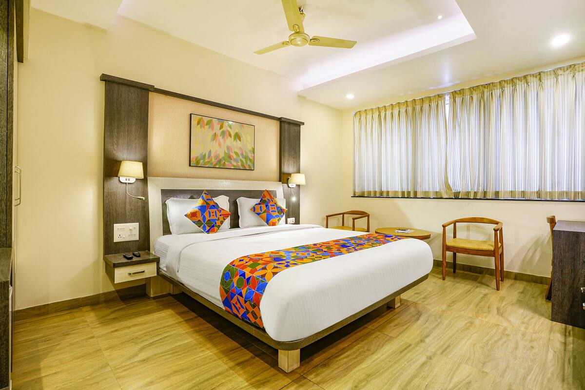 A neatly arranged hotel room with a large bed, a ceiling fan, a window with curtains, and a small wooden chair.
