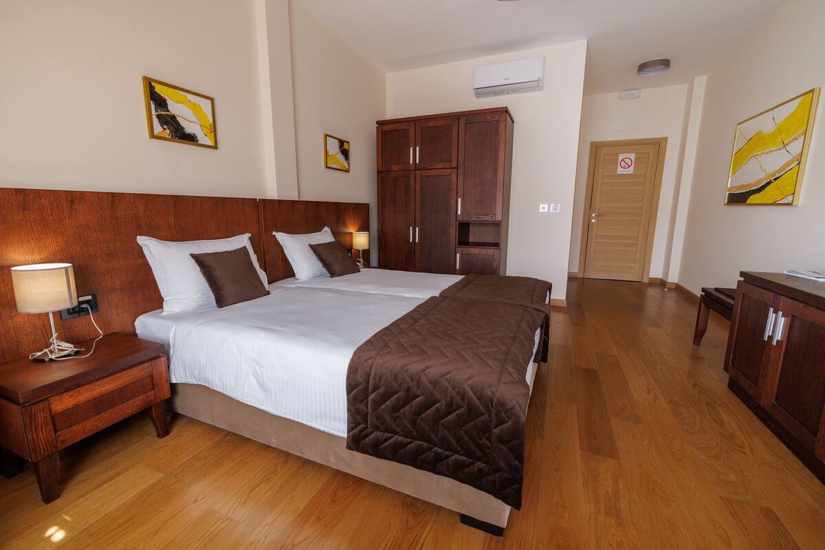 Una habitación de hotel con una cama grande, cabecera de madera, una mesita de noche con lámpara, un armario y obras de arte en las paredes.
