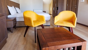 Une chambre d’hôtel avec un plancher en bois, une table en bois, deux chaises jaunes, un lit avec une literie blanche, une armoire en bois et un tableau accroché au mur.