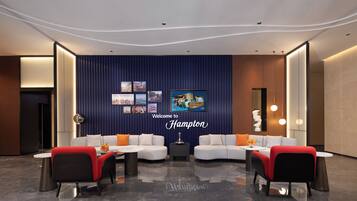 A modern lobby with a curved ceiling, a reception desk, two sofas with red and black chairs, and a wall with framed pictures and a 'Welcome to Hampton' sign.