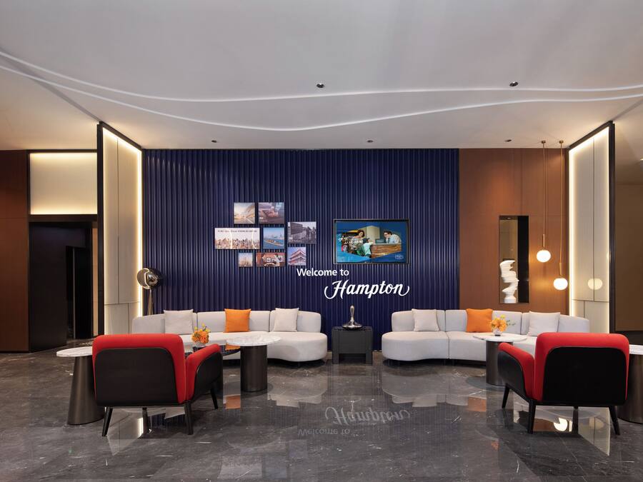 A modern lobby with a curved ceiling, a reception desk, two sofas with red and black chairs, and a wall with framed pictures and a 'Welcome to Hampton' sign.