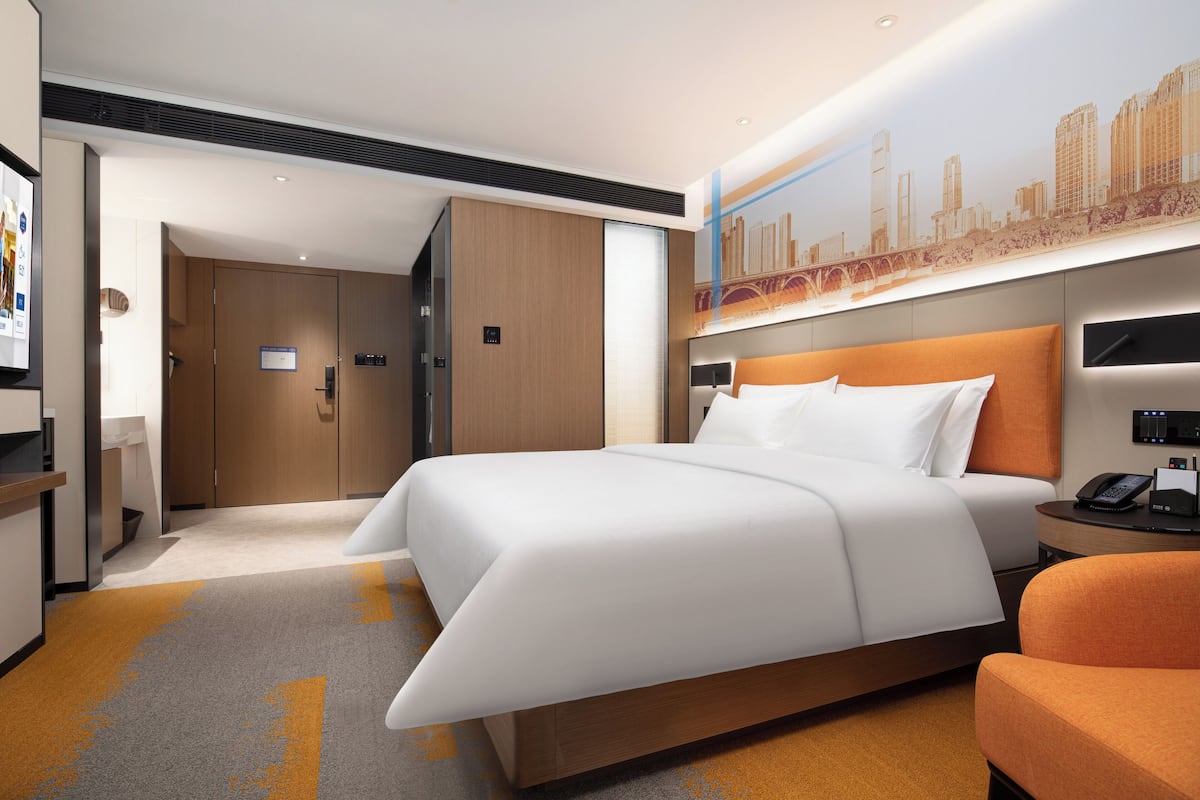A modern hotel room with a large bed, a cityscape mural, a wooden wardrobe, and a small orange chair.