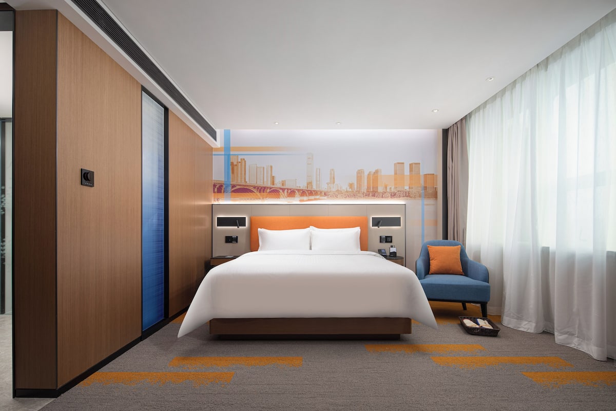 A modern hotel room with a large bed, a cityscape mural on the wall, a blue armchair, and a large window with sheer curtains.