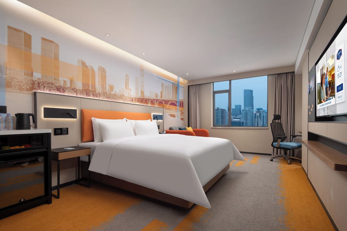 A modern hotel room with a large bed, a desk, a chair, and a city view.