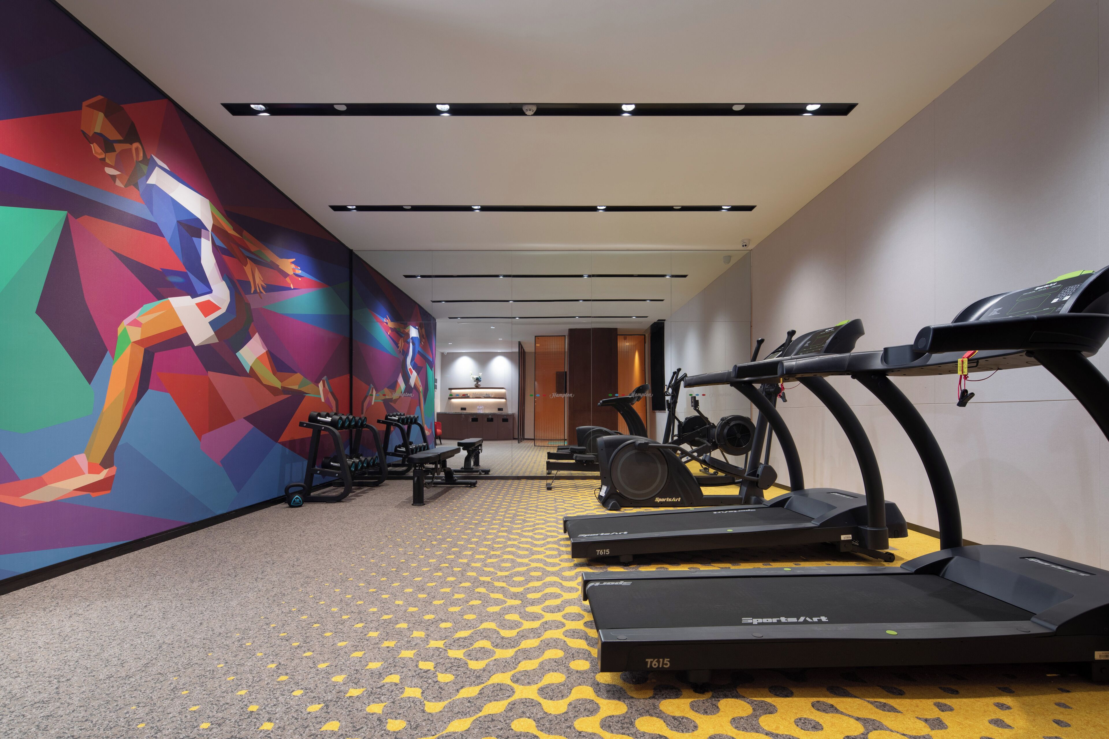 Un gimnasio moderno con cintas de correr, bicicletas estáticas y un mural colorido en la pared.