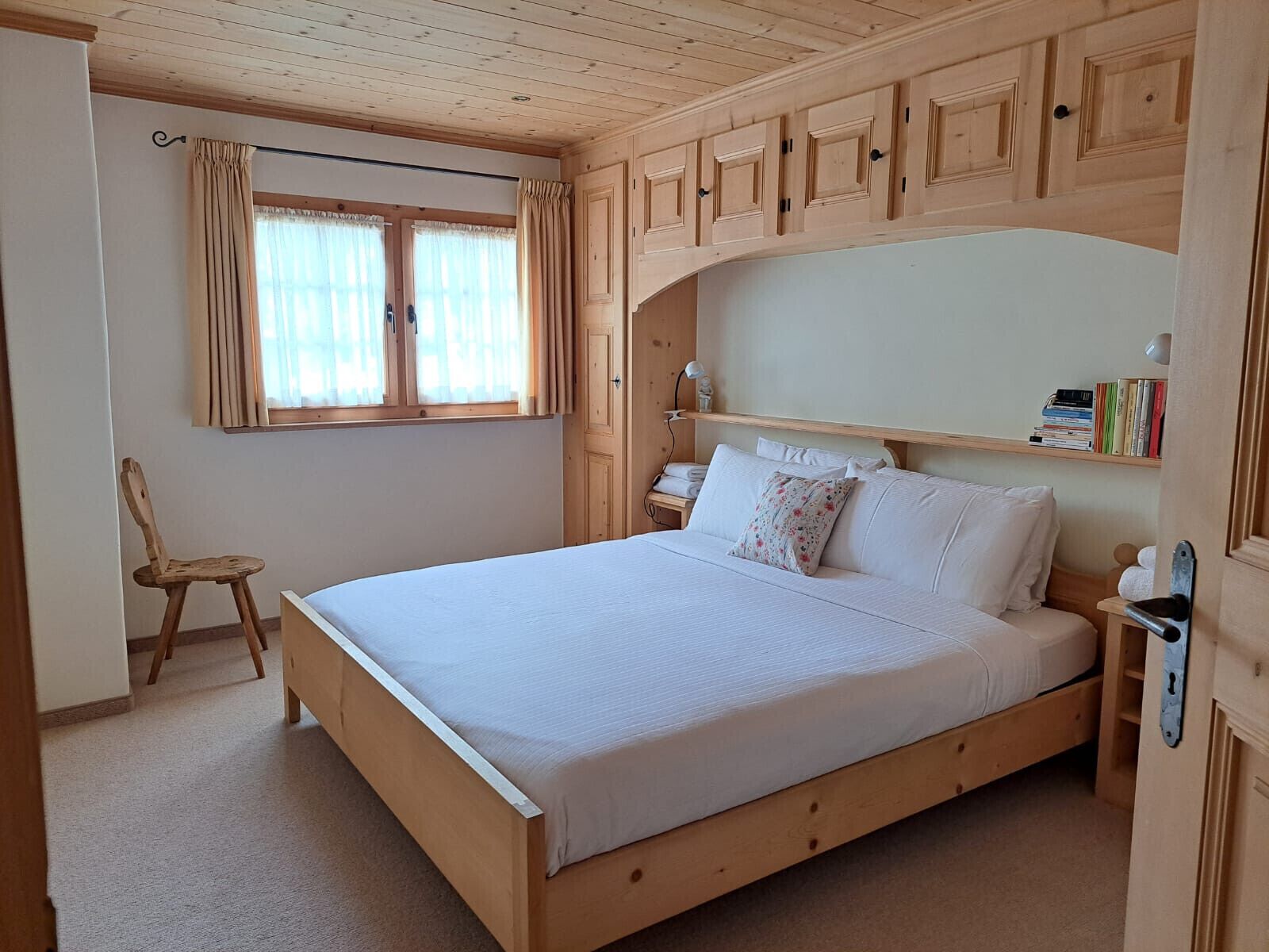 A wooden-framed bedroom with a bed, a chair, a nightstand, and a shelf with books.