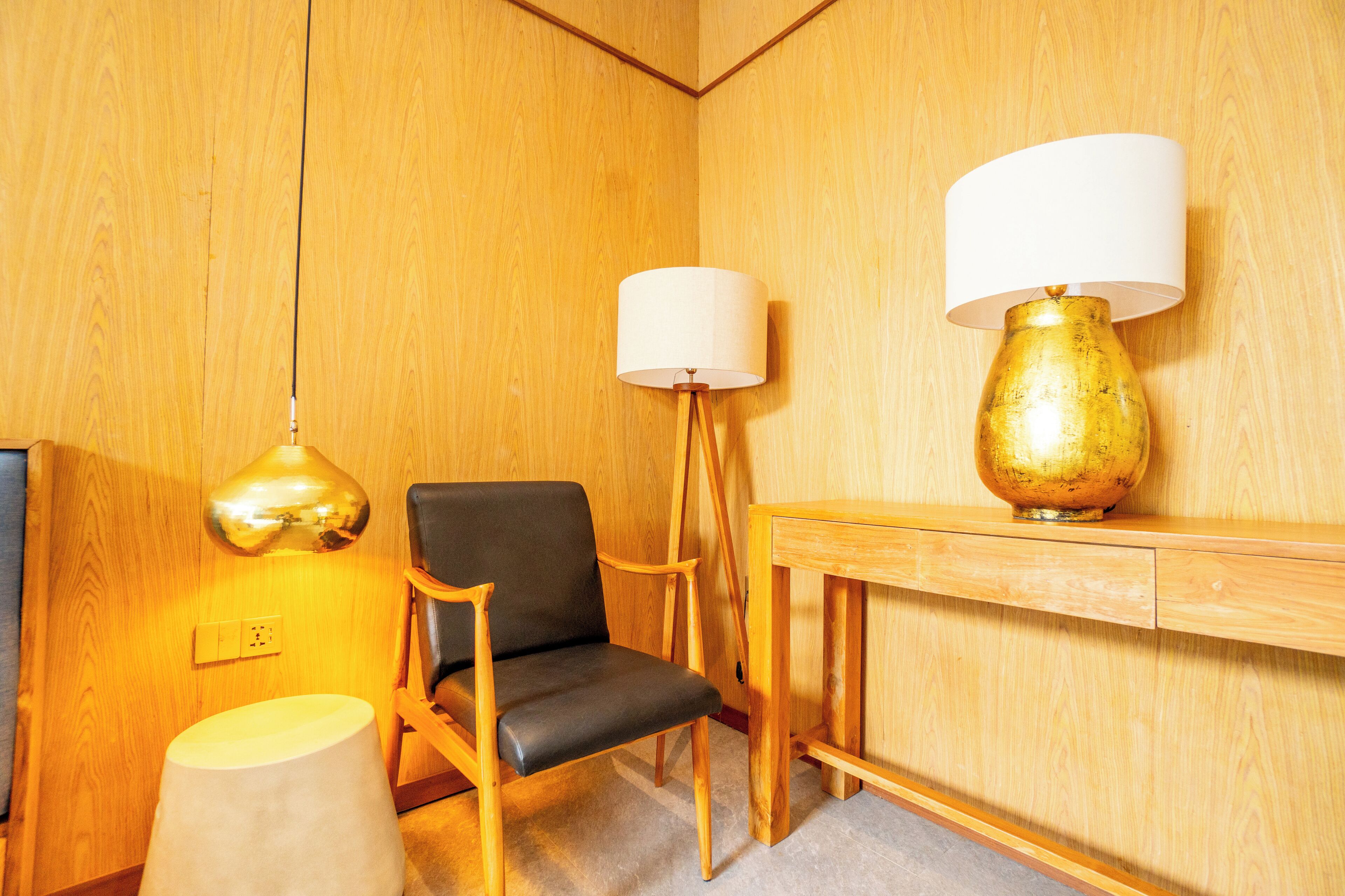 Una habitación con paredes de madera, una silla negra, una mesa de madera con una lámpara dorada y una lámpara blanca.