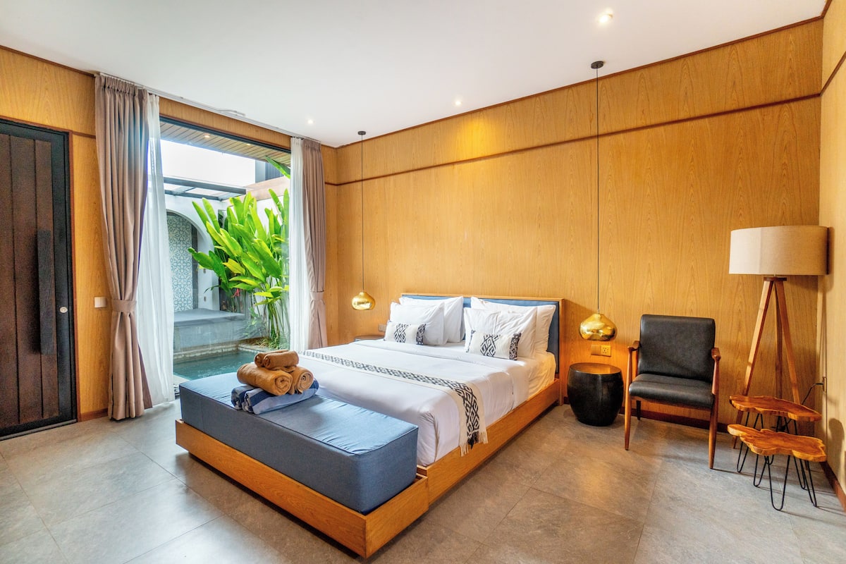 A modern bedroom with a large bed, a seating area, a lamp, and a view of a pool.