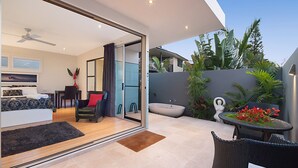 A modern hotel room with a sliding glass door leading to a balcony. The room includes a bed, a desk, and a chair. The balcony has a table and chairs, and there are plants and a decorative sculpture.
