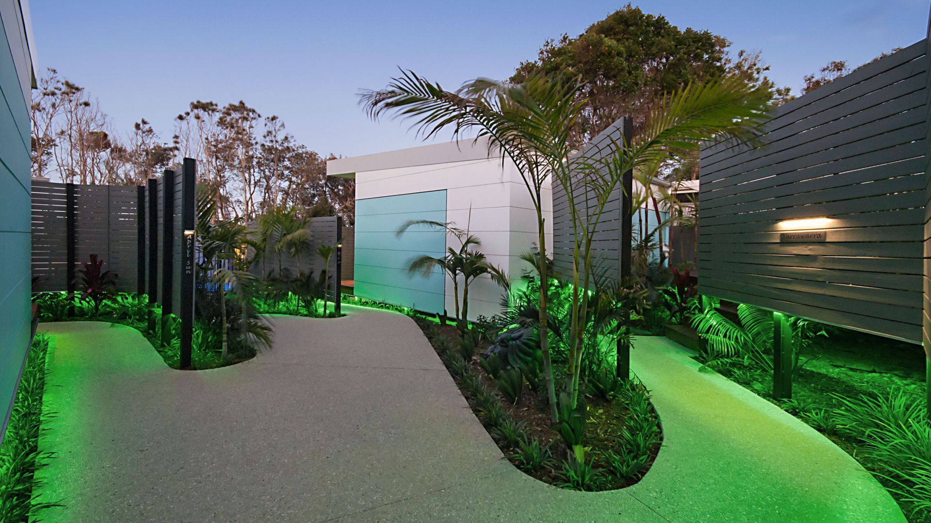 A modern building with a curved pathway, surrounded by tropical plants and palm trees.