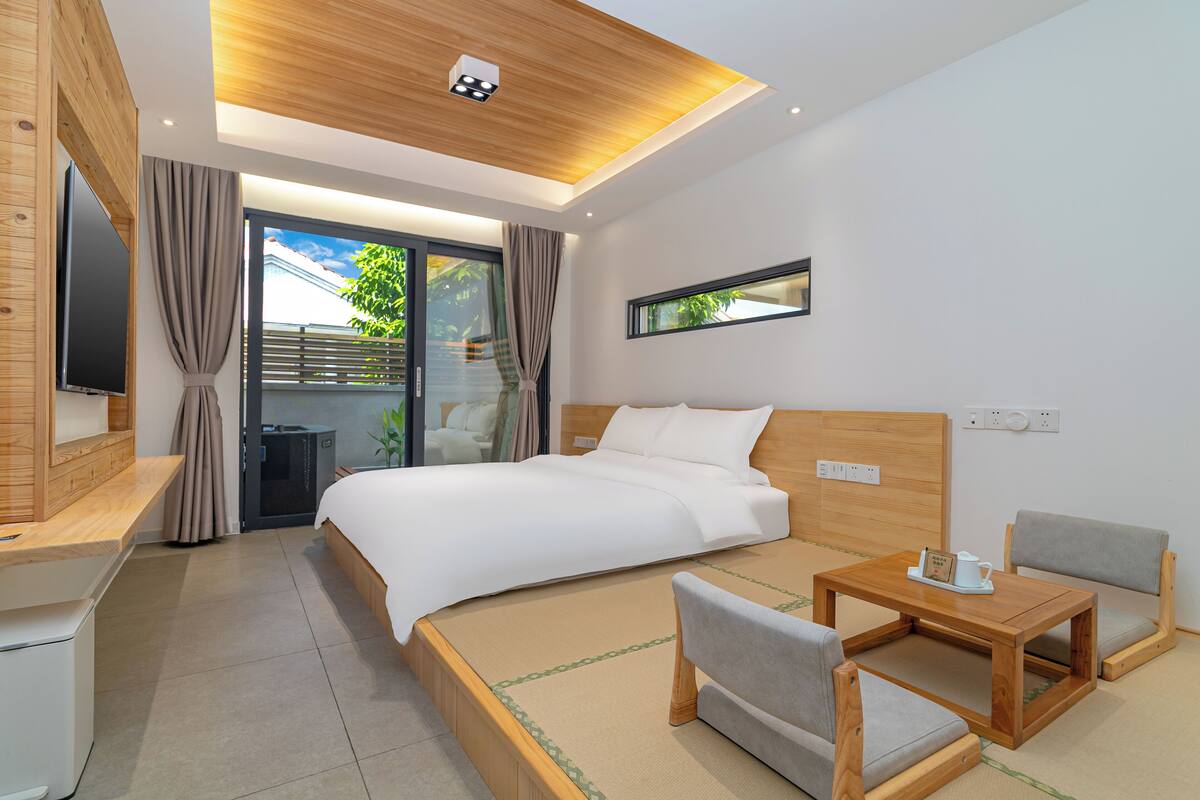 Ein modernes Hotelzimmer mit einem großen Bett, einem kleinen Holztisch, zwei Stühlen, einem Fernseher und einem Fenster mit Blick auf Grünflächen.