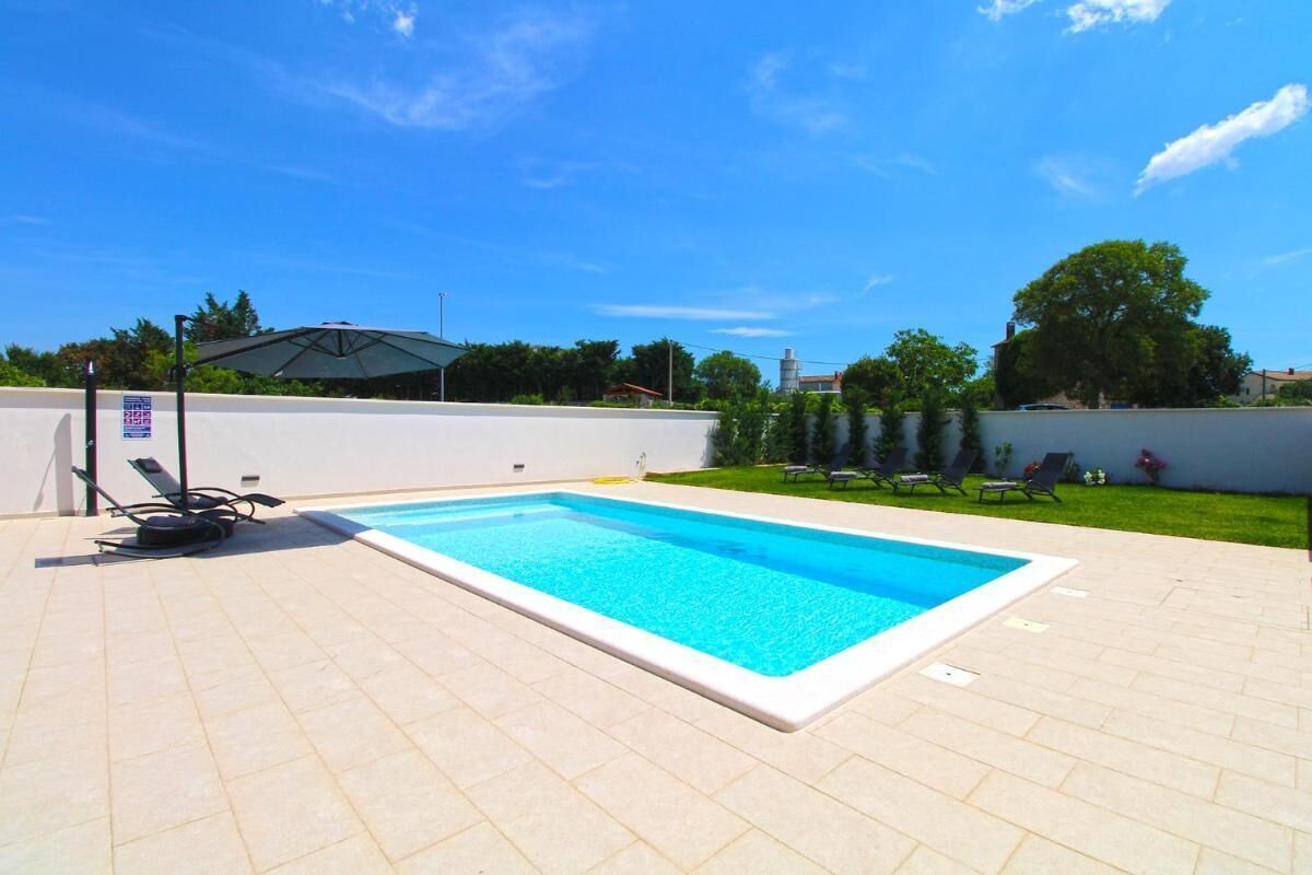 A rectangular swimming pool with a white edge, surrounded by a tiled patio. There are lounge chairs and a table with umbrellas in the background.