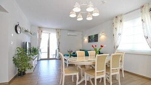 Een eetgedeelte met een ronde tafel, stoelen en een kroonluchter. Achtergrond toont een bank en een raam met gordijnen.