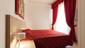 Une petite chambre avec un lit simple, une tête de lit en bois, une table de chevet avec une lampe et des rideaux rouges.