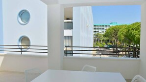 Ein moderner Außenbereich zum Sitzen mit einem weißen Tisch und Stühlen, einem Balkon mit Geländer und Blick auf ein Gebäude sowie Bäume im Hintergrund.