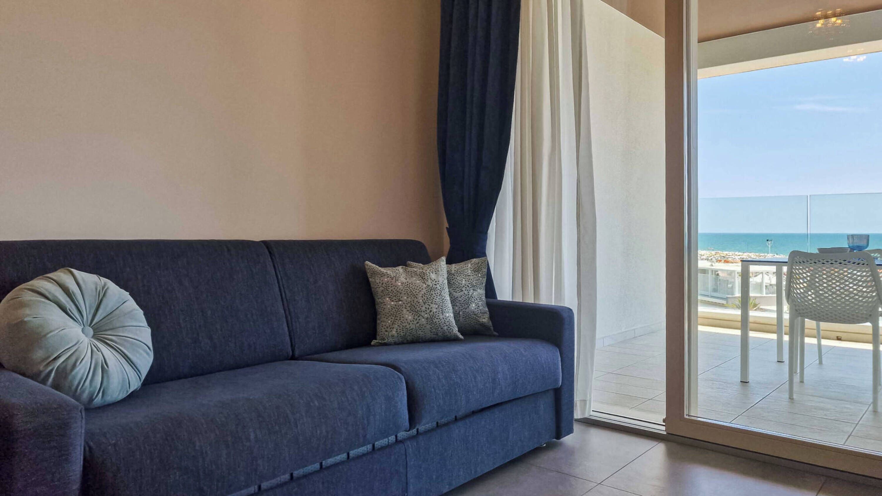 A dark blue sofa with two patterned pillows, a glass door leading to a balcony with a view of the sea.