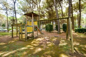 Un espace de jeux avec une structure d’escalade en bois et des balançoires, situé dans un parc arboré.