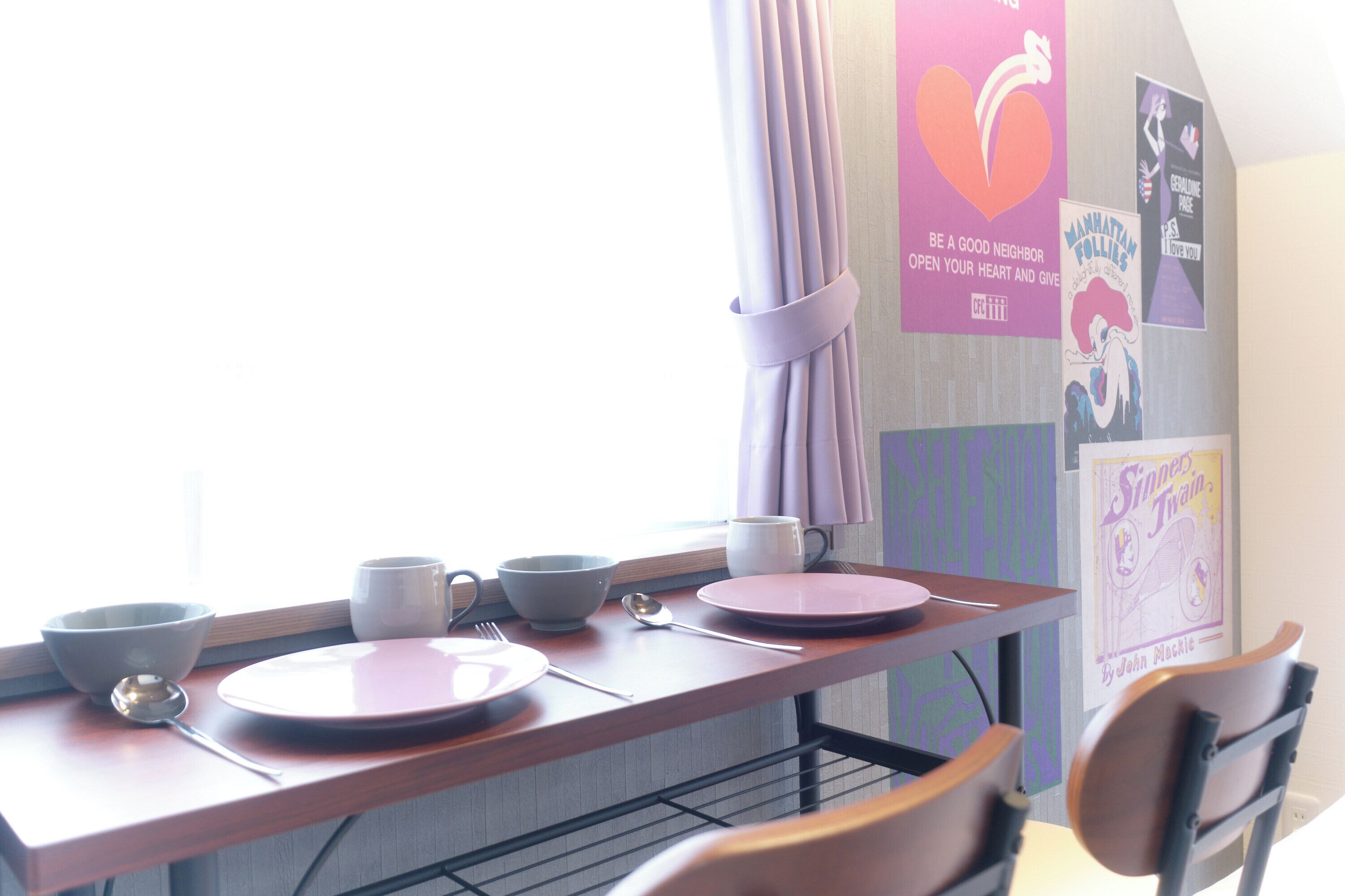 A dining area with a table set for two, including plates, bowls, cups, and cutlery. There are posters on the wall and a window with curtains.
