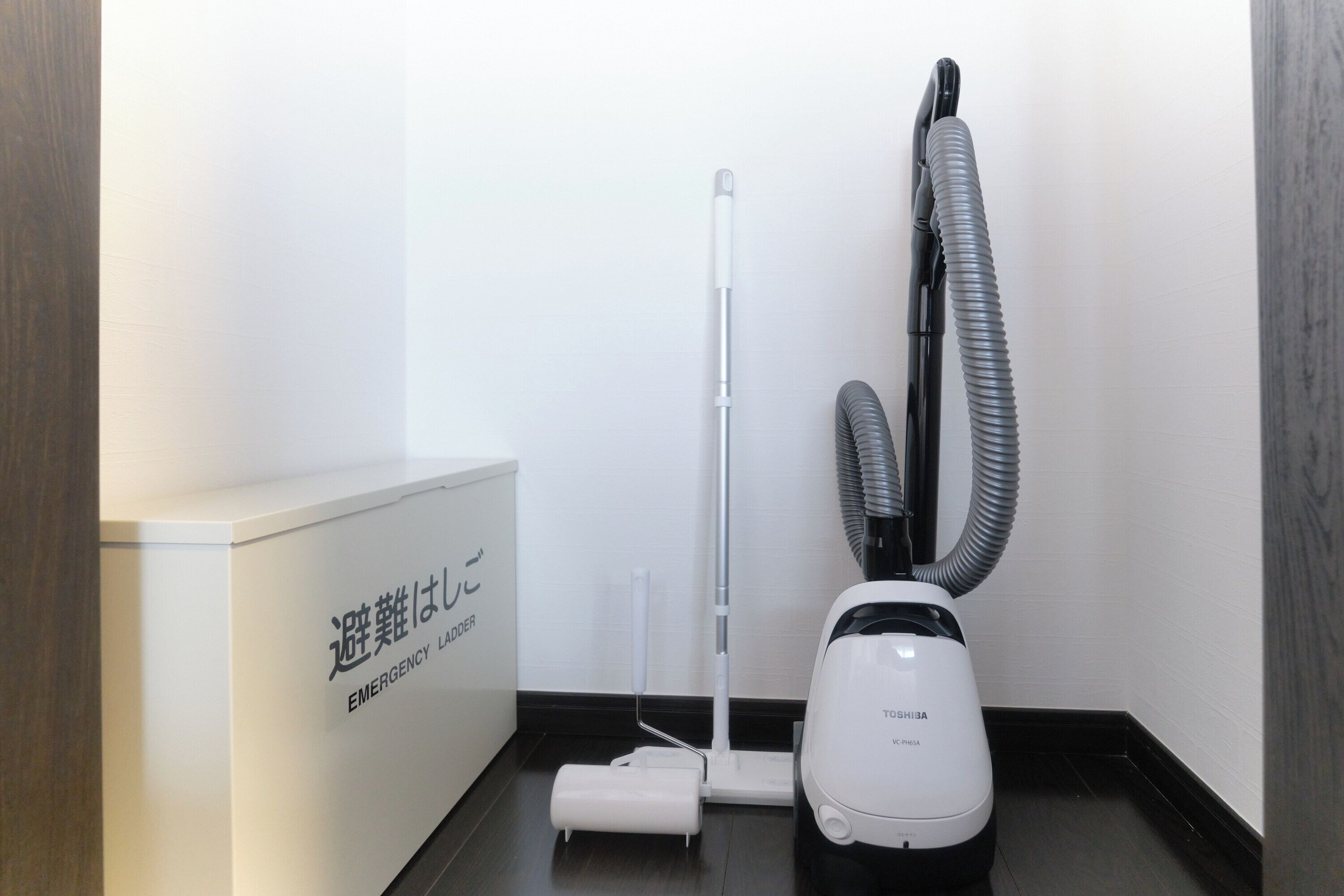 A corner of a room with a white emergency ladder, a white vacuum cleaner, and a black vacuum hose.