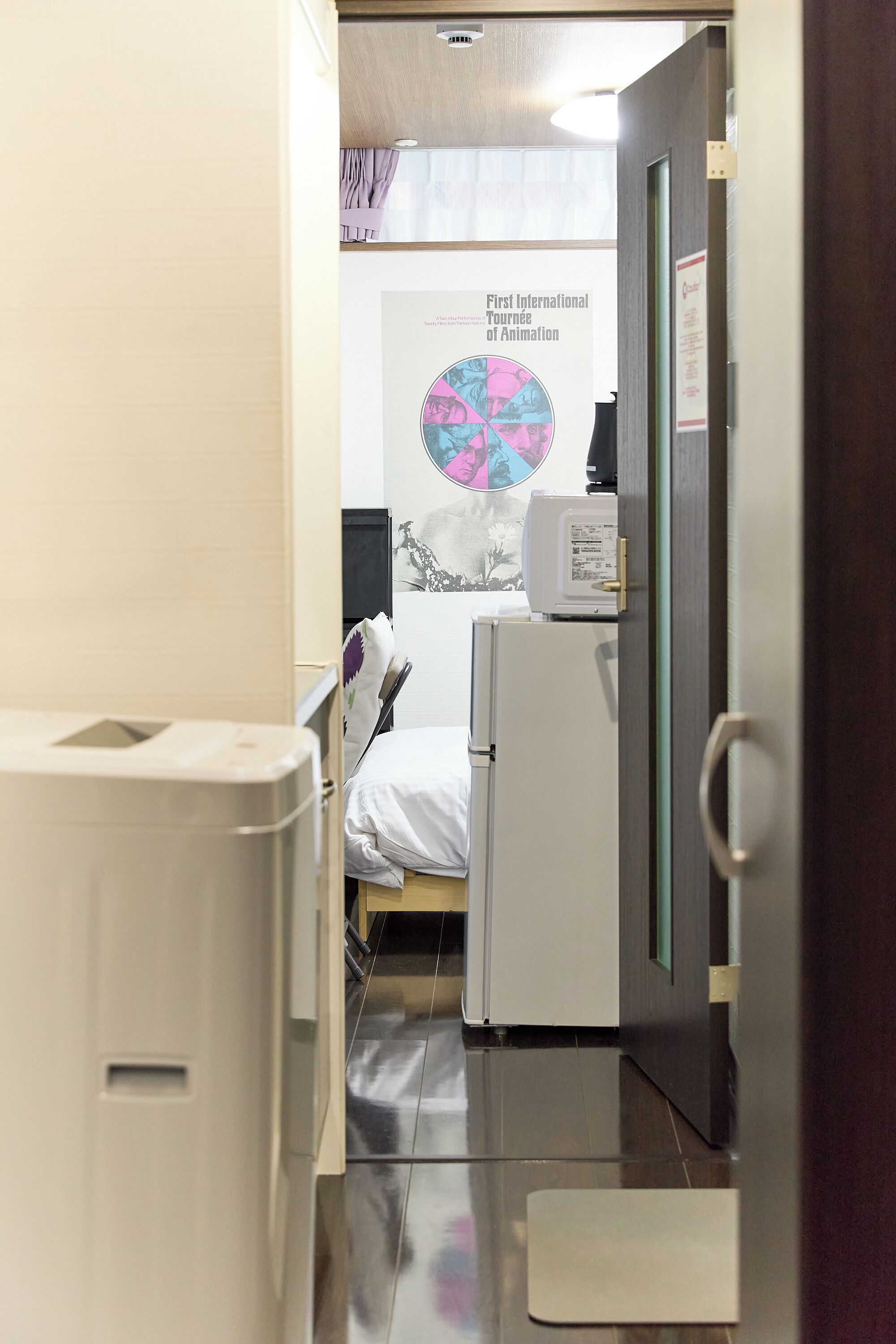 Un pasillo estrecho que conduce a un dormitorio con una cama, un refrigerador blanco pequeño y un cartel en la pared.