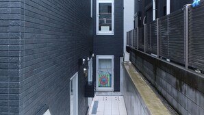 A narrow alleyway with a door featuring a colorful design, flanked by dark brick walls and a metal fence. - Marine Cove Hatsudai by Tranova (Tokyo)