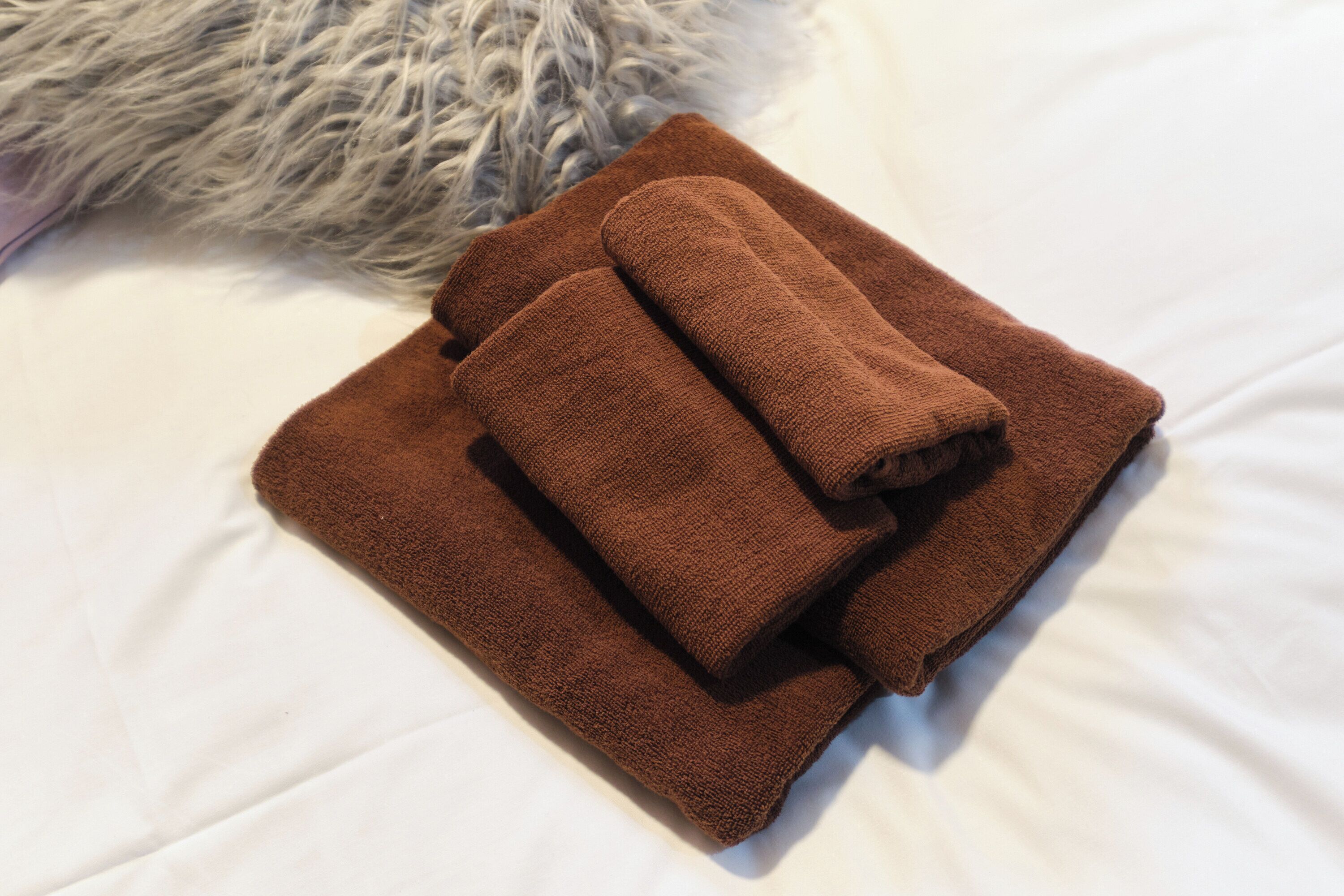 Una toalla marrón doblada con cuidado sobre una cama con sábanas blancas y un almohadón gris mullido.