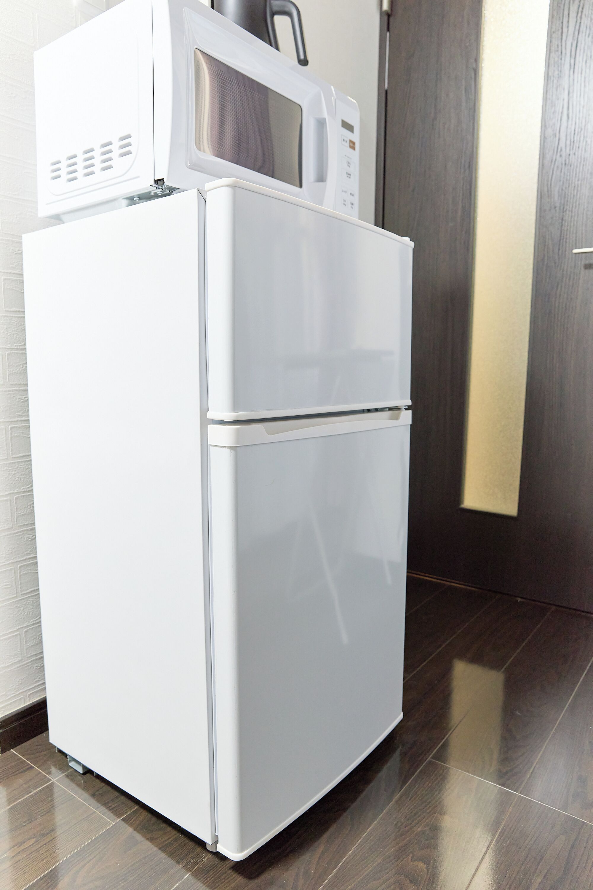 A white refrigerator with a microwave on top, located in a room with a wooden floor and a door with a gold accent.